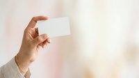 Person with medium skin tone holding blank white card or business card against blurred background for branding, marketing, or corporate identity mockup purposes.