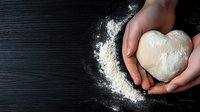 Heart shaped raw dough in female hands on black wooden background with scattered flour, top view. Baking and cooking concept for Valentine's day or love theme.