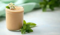 Creamy beige smoothie in glass jar decorated with white flowers and fresh mint leaves on light background with soft bokeh effect and copy space.