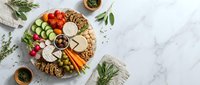 Gourmet cheese and charcuterie board with crackers, nuts, vegetables and herbs arranged on marble surface for entertaining and appetizers.