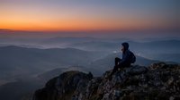 Silhouetted male hiker sitting on rocky mountain peak at sunrise, overlooking layered misty valleys and distant mountains. Contemplative outdoor adventure and travel concept.