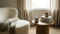 Cozy reading corner with cream armchair, knitted throw blanket, wooden side table, coffee cup and books by sunny window with natural light streaming in.