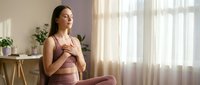 Young pregnant woman practicing mindfulness meditation at home, sitting peacefully in lotus position with hands on belly in bright living room.