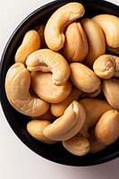 Fresh roasted cashew nuts in black ceramic bowl on white background, macro food photography showing detailed texture and natural golden color.
