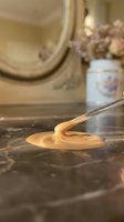 Creamy peanut butter dripping from wooden spoon onto dark marble countertop with blurred kitchen background and dried flowers in jar.