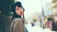 Young Asian woman with headphones listening to music on urban street with bokeh background and warm sunlight creating dreamy atmosphere for lifestyle content.