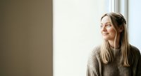 Young woman with long brown hair and fair complexion, wearing a beige knit sweater, gazing thoughtfully out a window with natural light. Portrait suitable for lifestyle, wellness, and editorial use.