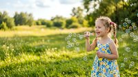 Young girl with blonde hair in braids, wearing a colorful floral dress, blowing bubbles outdoors in a sunny meadow surrounded by green grass and trees.