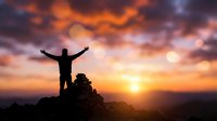 Man silhouette with raised arms celebrating success on mountain peak at dramatic sunset with orange and purple clouds in sky for motivation concepts.