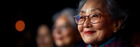 Senior Asian woman with glasses smiling warmly in soft lighting, representing wisdom, experience and positive aging for healthcare and lifestyle content.
