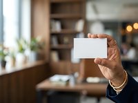 Close-up of hand holding blank white card in modern office workspace with wooden furniture and shelving, suitable for business branding and corporate messaging.