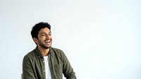 Young man with dark curly hair and olive skin tone, wearing olive green jacket over white shirt, smiling while looking to the side against white background for portrait and lifestyle use.