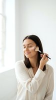 Young Hispanic woman applying makeup with brush in bright bathroom wearing white robe. Morning beauty routine skincare cosmetics preparation lifestyle.
