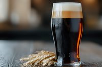 Glass of dark craft beer with creamy foam head and golden wheat ears on rustic wooden table, selective focus highlighting rich amber-brown color and fresh pour.