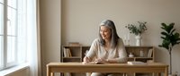 Senior woman with gray hair and light skin tone, wearing a beige cardigan, sitting at a wooden desk in a bright, minimalist home office with plants and shelving, working on a laptop for remote work or freelance projects.