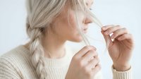 Senior woman with long gray braided hair wearing cream knit sweater, touching her face with both hands in contemplative pose against white background for wellness and aging content.