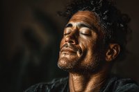Middle-aged Hispanic man with closed eyes and peaceful expression enjoying refreshing water splash on face in dramatic lighting for wellness concepts.