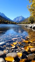Tranquil mountain river with colorful stones reflecting autumn sunlight, snow-capped peaks and yellow trees along shoreline against clear blue sky.