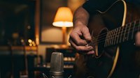 Musician playing acoustic guitar in cozy home recording studio with vintage microphone and warm ambient lighting for music production and songwriting.