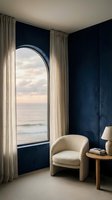 Modern coastal bedroom corner with cream armchair, wooden side table, and arched window overlooking serene ocean view, navy walls, beige curtains, contemporary interior design.