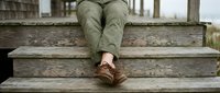 Person in khaki pants and brown shoes sitting on weathered wooden steps of rustic building, casual outdoor lifestyle and relaxation concept.