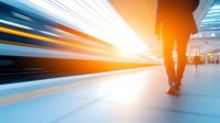 Blurred motion of fast moving train at station platform with silhouette of walking person, dramatic orange sunset light creating dynamic urban scene.