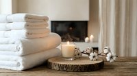 Spa relaxation setup with white towels, candle and cotton flowers on wooden tray near fireplace creating peaceful wellness atmosphere for home comfort.