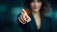 Confident businesswoman in dark suit pointing finger forward, demonstrating leadership and decision-making in corporate environment.