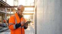 Construction worker in orange safety vest using digital tablet at building site during golden hour for project management and quality control.