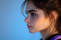 Young Caucasian woman in profile view against blue background, dramatic side lighting highlights facial features and loose strands of dark hair in messy updo hairstyle.