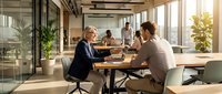 Professional businesswoman and businessman shaking hands across conference table in modern office meeting room with colleagues in background.