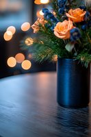 Peach roses and blue flowers in black vase with pine branches against blurred bokeh lights creating cozy winter atmosphere on wooden table surface.
