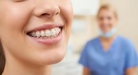 Close-up of young woman's bright smile showing straight white teeth with dental braces at orthodontist office with female dentist in background.