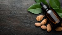 Amber glass bottle with dropper surrounded by fresh almonds and green leaves on dark slate background for natural skincare and aromatherapy products.