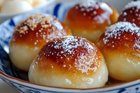 Sweet buns with golden brown tops dusted with powdered sugar and chopped nuts served in traditional blue and white ceramic bowl, fresh bakery close-up.