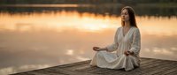 Young woman meditating in lotus pose on wooden dock at sunset by calm lake. Peaceful mindfulness practice for wellness and mental health concepts.