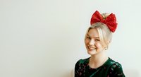 Young woman with blonde hair and fair complexion wearing a red sequined bow headband and dark green sparkly top, smiling against neutral background for holiday and festive celebrations.