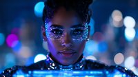 Young African American woman with glowing neon makeup and rhinestones around eyes in futuristic fashion setting with blue bokeh lights and sparkles in background.