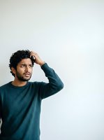 Young Middle Eastern man with curly dark hair wearing teal sweater, hand to head in thoughtful pose against white background. Suitable for lifestyle, emotion, and casual apparel marketing.