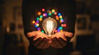 Hands holding glowing light bulb with colorful bokeh lights creating halo effect in dark room representing creativity innovation and bright ideas concept.