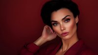 Young woman with short dark hair in burgundy outfit against deep red background, hand elegantly touching face with confident expression.