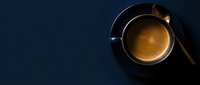 Overhead view of freshly brewed espresso in black cup with saucer and spoon on dark blue background, suitable for coffee shop menus and beverage marketing.