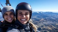 Two young women paragliding together over mountain landscape, smiling at camera during extreme sport adventure flight with scenic valley view below.
