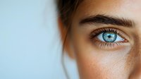Close-up macro shot of bright blue eye with natural freckles and long eyelashes against light background, showing natural beauty and unique facial features.