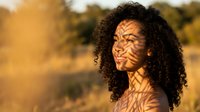 Young African American woman with curly hair enjoying warm sunlight filtering through leaves creating natural shadow patterns on her face during golden hour.