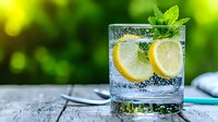 Refreshing glass of sparkling water with lemon slices and fresh mint leaves on rustic wooden surface against blurred green garden background. Cooling summer drink.