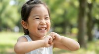 Happy Asian girl child smiling with hands clasped together in outdoor park setting. Joyful expression shows excitement and positive emotions for family lifestyle content.