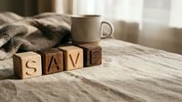 Wooden letter blocks spelling SAVE on a linen surface with a mug and cozy blanket in soft natural light, evoking home finance and savings concepts.