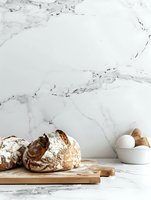 Fresh rustic bread loaves dusted with flour on wooden cutting board against marble background, with eggs in ceramic bowl, minimalist food styling composition.