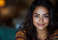 Young woman with dark wavy hair and natural makeup smiling warmly at camera. Soft bokeh background creates intimate portrait atmosphere with teal and amber tones.